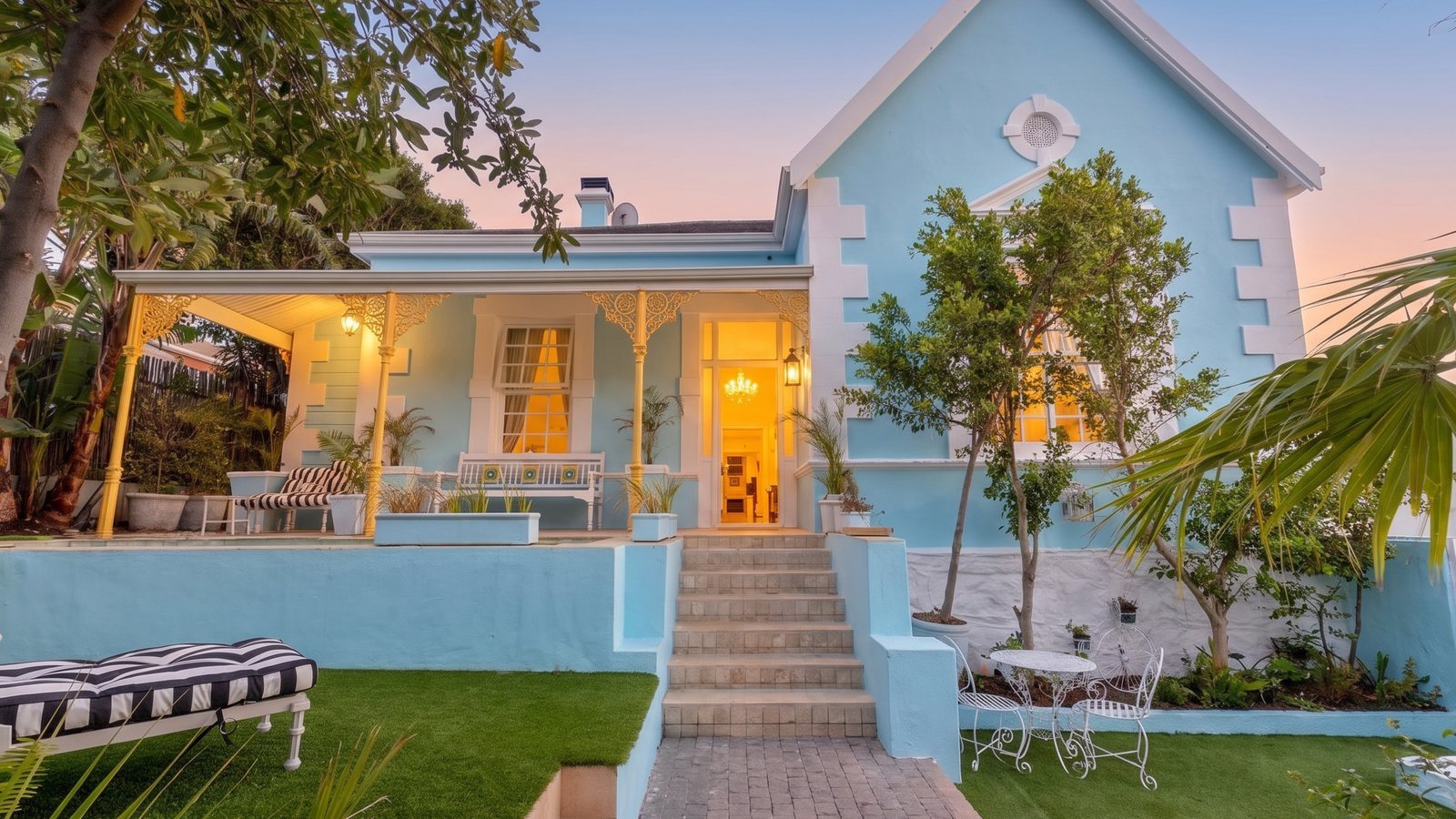 The front of Amina at dusk: a powder-blue house with a yellow lace verandah, the front door open and the chandelier lit