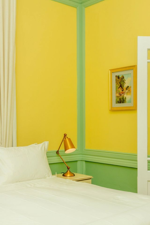 A queen bed against the yellow and green wall, a reading lamp beside it